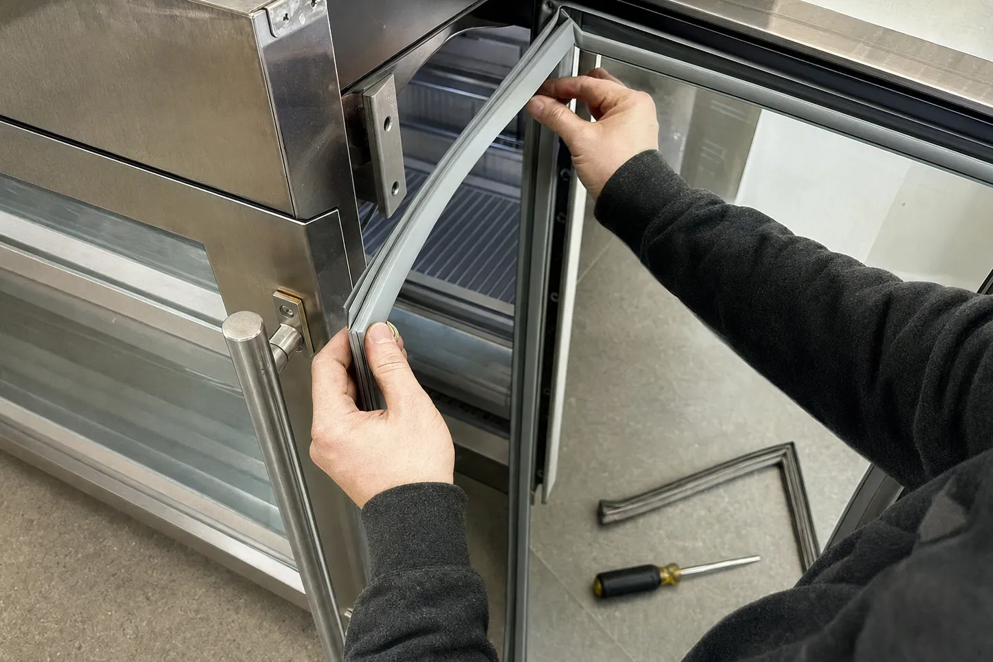 Hands pressing a new grey door gasket into the channel of an open stainless door, the old seal on the floor