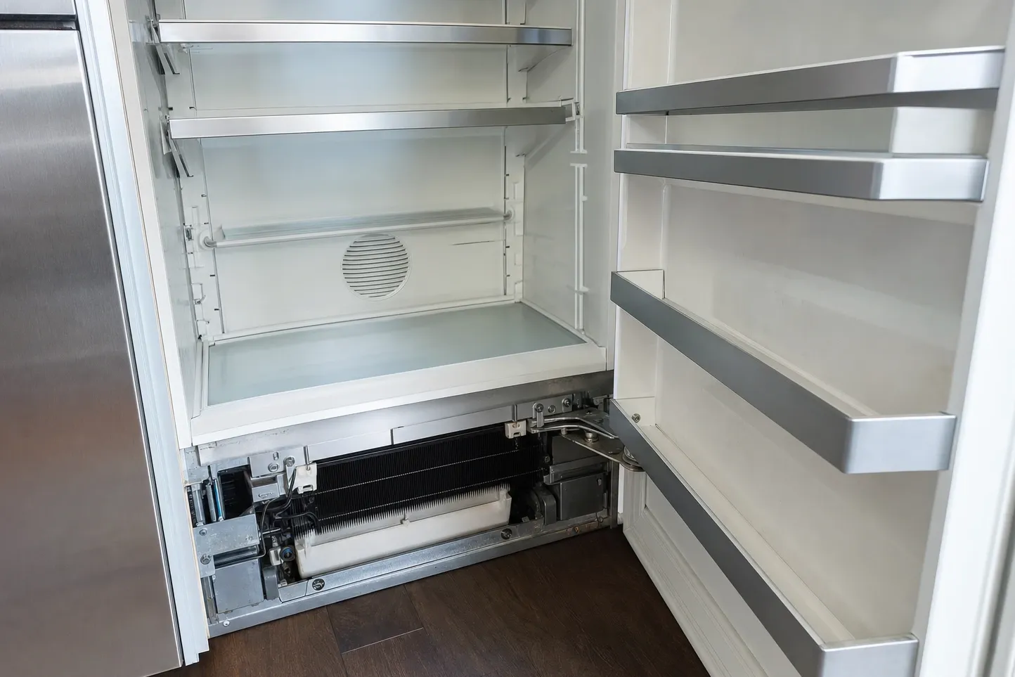 Empty built-in refrigerator interior with the base panel removed, exposing the finned evaporator coil and drain tray